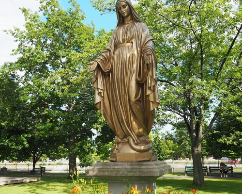Goldene Statue der Jungfrau Maria mit offenen Armen auf einem Sockel, umgeben von Blumen und Bäumen in einer gepflegten Gartenanlage bei blauem Himmel.