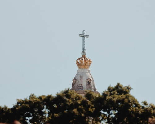 Nahaufnahme der mit einer goldenen Krone verzierten Kuppel der Basilika von Fátima, überragt von einem Kreuz, im Vordergrund Baumwipfel.