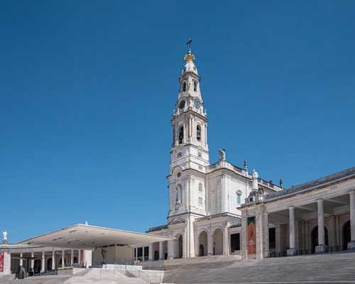 Seitliche Ansicht der Basilika Nossa Senhora do Rosário in Fátima mit hohem Glockenturm, angrenzendem Säulengang und großem überdachten Altarbereich vor strahlend blauem Himmel.