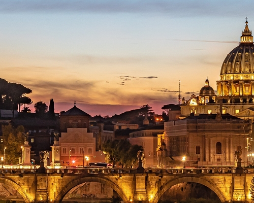 Abendlicher Blick auf den beleuchteten Petersdom und die Engelsbrücke in Rom, mit goldenem Licht auf der Kuppel vor farbenfrohem Himmel und spiegelndem Tiberfluss im Vordergrund.