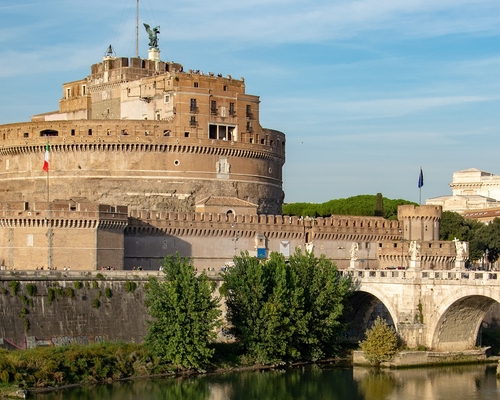 Blick auf die runde Engelsburg am Tiberufer in Rom, mit der historischen Engelsbrücke im Vordergrund und ruhigem Wasser, eingefasst von Bäumen und Gebäuden.