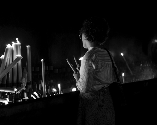 Eine Frau steht im Kerzenraum des Heiligtums von Fátima, hält mehrere Kerzen in der Hand und blickt auf brennende Kerzen in einer dunklen, stillen Atmosphäre.