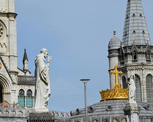 Nahaufnahme der Türme der Basilika von Lourdes mit der goldenen Krone und einem Kreuz auf der Kuppel, flankiert von Heiligenstatuen und architektonischen Verzierungen.